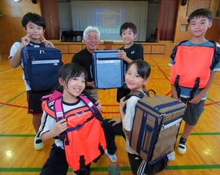 児童たちの声も参考に完成させた改良版の「ライフボックス」を手渡す細川晋さん（後列左から２人目）＝三重県鳥羽市堅神町、鳥羽小学校