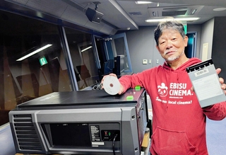 新たに導入した機材の前で、左手に新作のデジタルシネマデータ、右手に旧来のブルーレイディスクを持つ近兼さん＝メガヱビス。