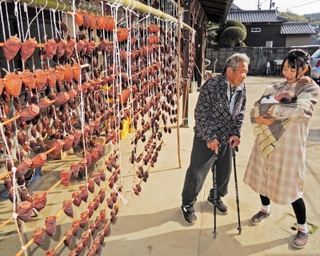 正月用「串柿」など干し柿のカーテンの前で、ひ孫を抱いた孫の高島依里さん（右）と談笑する田中秋雄さん＝加古川市志方町岡