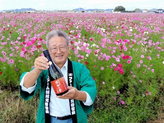 コスモス畑を背に、６年ぶりに醸した清酒「純米吟醸　稲加乃誉」を持つ「ファーム稲加　見谷営農」の小山和彦組合長＝稲美町加古