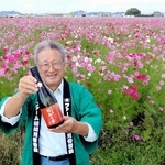 コスモス畑を背に、６年ぶりに醸した清酒「純米吟醸　稲加乃誉」を持つ「ファーム稲加　見谷営農」の小山和彦組合長＝稲美町加古