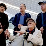 　米国への出発前、取材に応じる被爆者団体「長崎県被爆者手帳友の会」の代表団のメンバー。中央奥は朝長万左男さん＝２７日午前、羽田空港