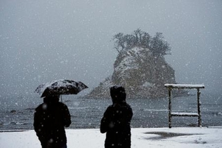 　雪が降りしきる中、石川県珠洲市の景勝地「見附島」を訪れた人たち。能登半島地震の発生から２年となった＝１日午前７時５分