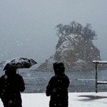 　雪が降りしきる中、石川県珠洲市の景勝地「見附島」を訪れた人たち。能登半島地震の発生から２年となった＝１日午前７時５分