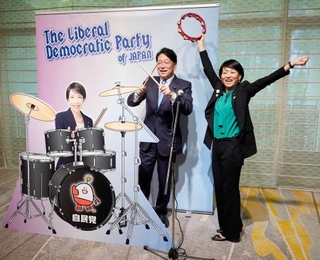 　自民党大会の会場に設置された高市首相のフォトパネルの横でポーズをとる小野寺五典税調会長（左）と鈴木貴子広報本部長＝１２日午前、東京都内