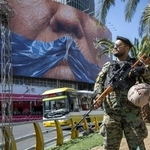 　イランの首都テヘランで、ホルムズ海峡の形をした青いリボンで口をふさがれたトランプ米大統領のポスターの前に立つ警察官＝５月（ゲッティ＝共同）