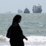 　ホルムズ海峡に臨む海岸を歩く女性。沖合には船舶が浮かんでいる＝２５日、イラン南部バンダルアバス（ＷＡＮＡ提供、ロイター＝共同）