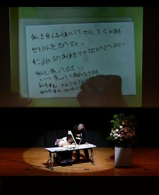 　新潟水俣病の公式確認から６０年となり、新潟市で開かれた式典。壇上左はメッセージを書く胎児性水俣病患者の古山知恵子さん＝２０２５年５月３１日午後