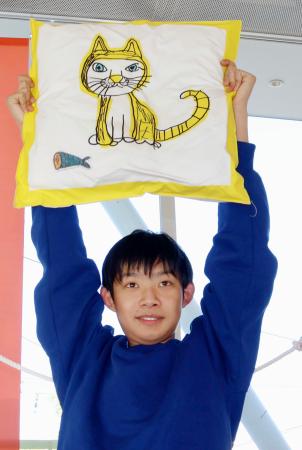 　自身が描いたネコがプリントされたクッションを掲げるぽんちゃん＝２０２５年１１月、前橋市