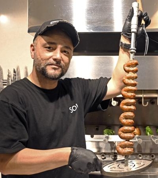 　母国ブラジルから取り寄せたシュラスコの専用装置（奥）で焼いた肉を持つ鈴木ウリセスさん＝北海道石狩市