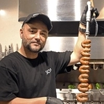 　母国ブラジルから取り寄せたシュラスコの専用装置（奥）で焼いた肉を持つ鈴木ウリセスさん＝北海道石狩市