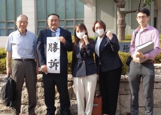 　判決後、代理人弁護士（両端）と共に「勝訴」と書いた紙を掲げる朝倉隆介さんら３人＝７月３０日、堺市