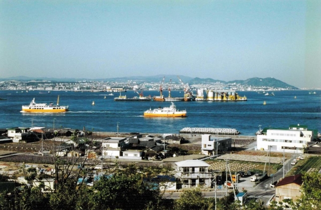 まだ大橋の姿がない明石海峡。２本の主塔が立つ地点にコンクリートプラント船が停泊し、基礎工事が進む（１９９１年４月）