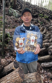 原木のそばで乾燥シイタケを手にする仲守さん＝猪名川町木間生