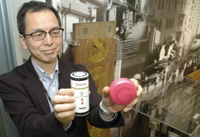 発売８０周年の丹頂チック（左）と国内シェア１位のワックス「ムービングラバー」を手にする藤原さん。後方にあるのは「丹頂マーク」のついた看板＝大阪市中央区十二軒町、マンダム本社