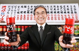 かつてお好み焼き店で実際に使われていた価格表の前で、「どろソース」を手にするオリバーソースの道満雅彦社長＝神戸市中央区港島南町３、同社本社