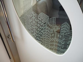 　京王９０００系のリニューアル車両で袖仕切りのガラスに描かれたイラスト。ビルの一つの屋上に天狗が立っている＝２０２５年３月８日、稲城市