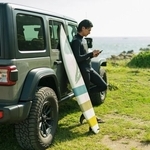 海辺の草原で車にサーフボードを立てかけ、波情報を確認するサーファー※画像はイメージです （One/stock.adobe.com）