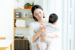 赤ちゃんを抱きながらスマートフォンで調べものをする母親 ※画像はイメージです（wheeljack/stock.adobe.com）
