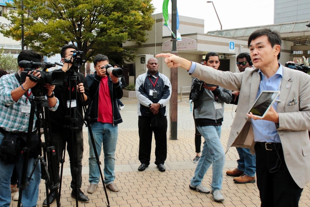 海外のテレビ局関係者にアプリで映像を見せながら震災当時の街の様子を説明する市職員の松崎太亮さん（右）＝神戸市長田区若松町４