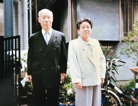 金婚式の式典に出席するため、正装に身を包む芝重吉さん（左）とヒデ子さん。右側の建物が阪神・淡路大震災で崩壊した自宅の文化住宅＝１９９１年４月、神戸市中央区筒井町３（家族提供）