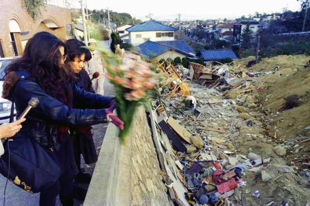 亡くなった友人を悼み、土砂崩れの現場へ花を投げ入れる女性ら。両脇から放送局のマイクが差し出されている＝１９日、西宮市仁川町６